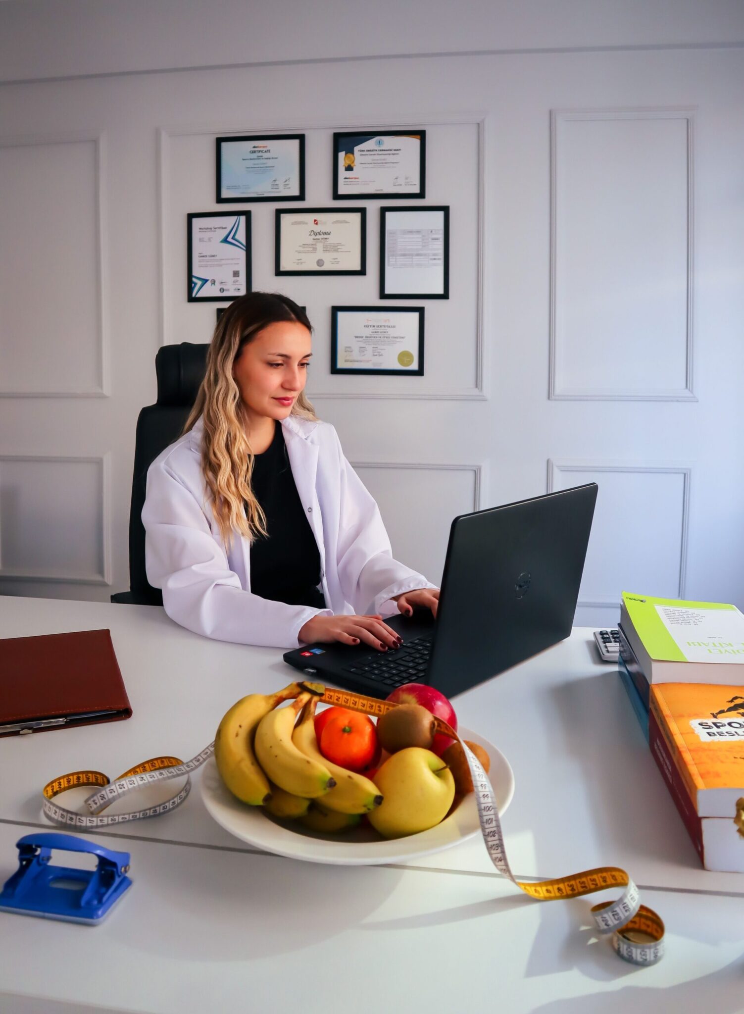 Female dietitian working on a laptop in a clinical office with nutrition-related books, fruits, and measuring tape on the desk representing professional nutrition consultation and healthy lifestyle planning